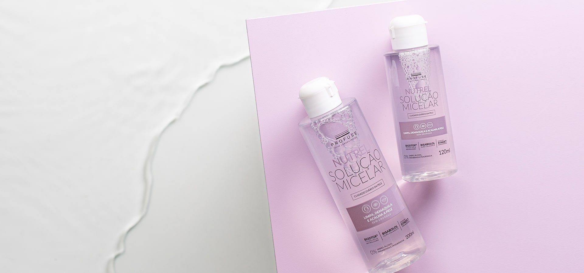 Uma embalagem de Nutrel Solução Micelar 120ml ao lado de outra de 200ml em cima de um cartão lilás. Ao fundo do cartão lilás, aparece um fundo branco com água.